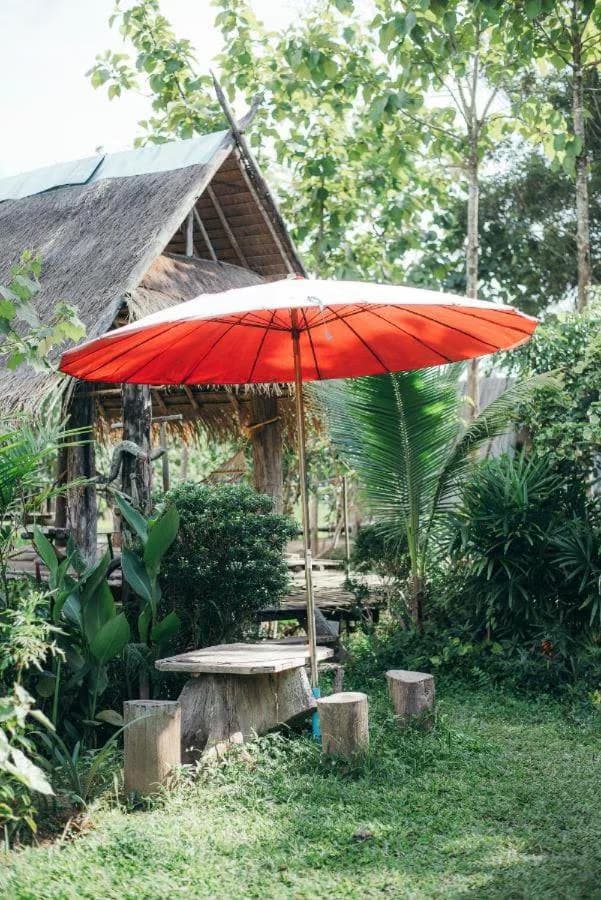 Camp View Mountain, Pai — red Thai parasol over a rustic log-stump table in a leafy garden beside the thatched bamboo huts