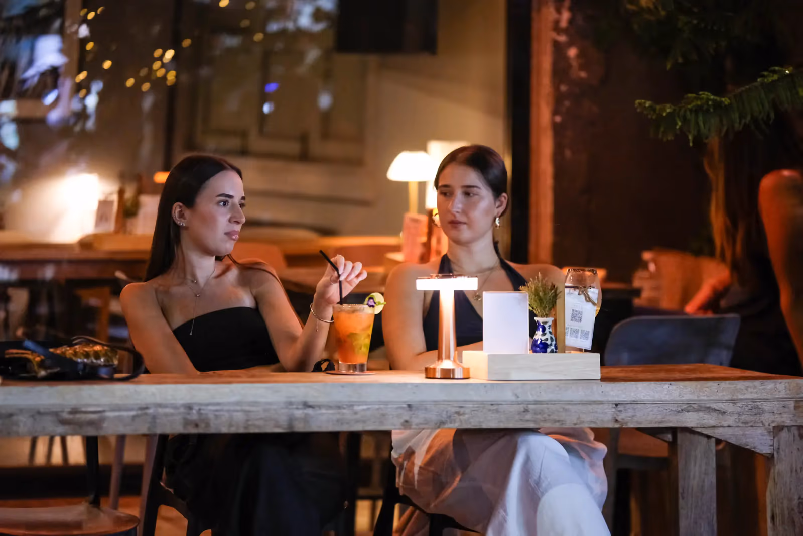 Two guests with cocktails at a lamp-lit front table at Vermut Café, the walking street behind the glass