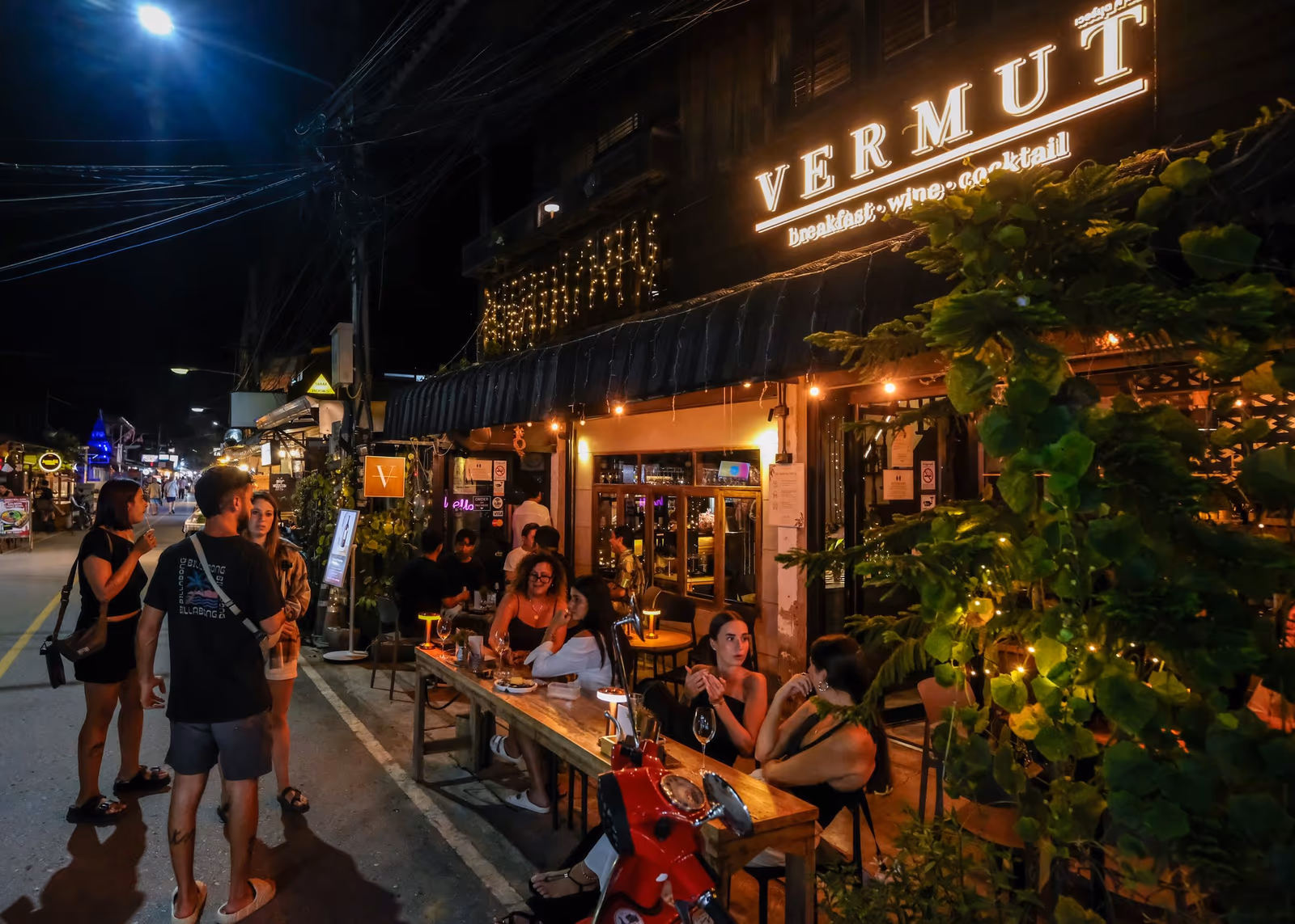 Vermut Café at night on Pai Walking Street — lamp-lit tables on the street, guests at the long table, the lit VERMUT sign above